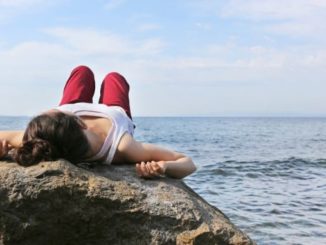Frau liegt meditierend auf einem Fels am Meer