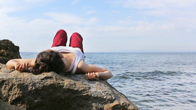 Frau liegt meditierend auf einem Fels am Meer
